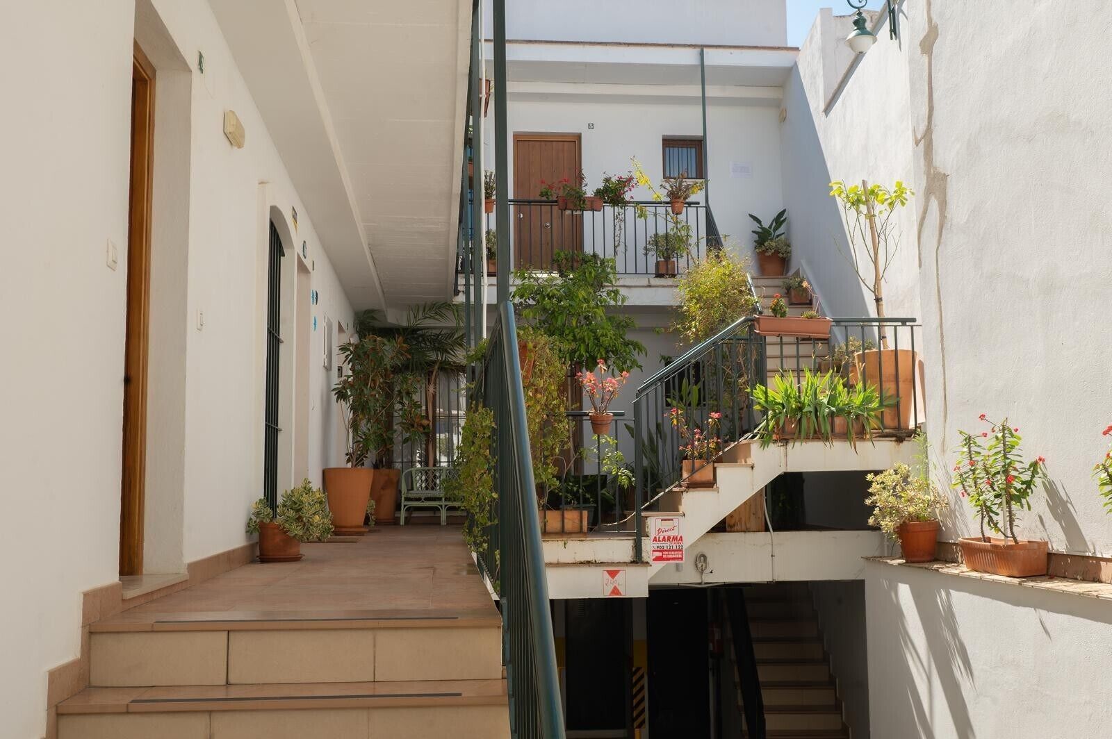Une cour étroite avec un escalier menant à un balcon du deuxième étage, entourée de plantes en pot et d’un banc.