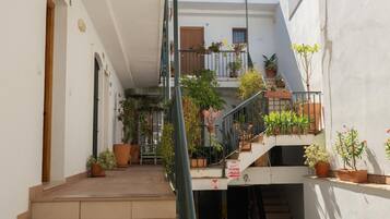Une cour étroite avec un escalier menant à un balcon du deuxième étage, entourée de plantes en pot et d’un banc.
