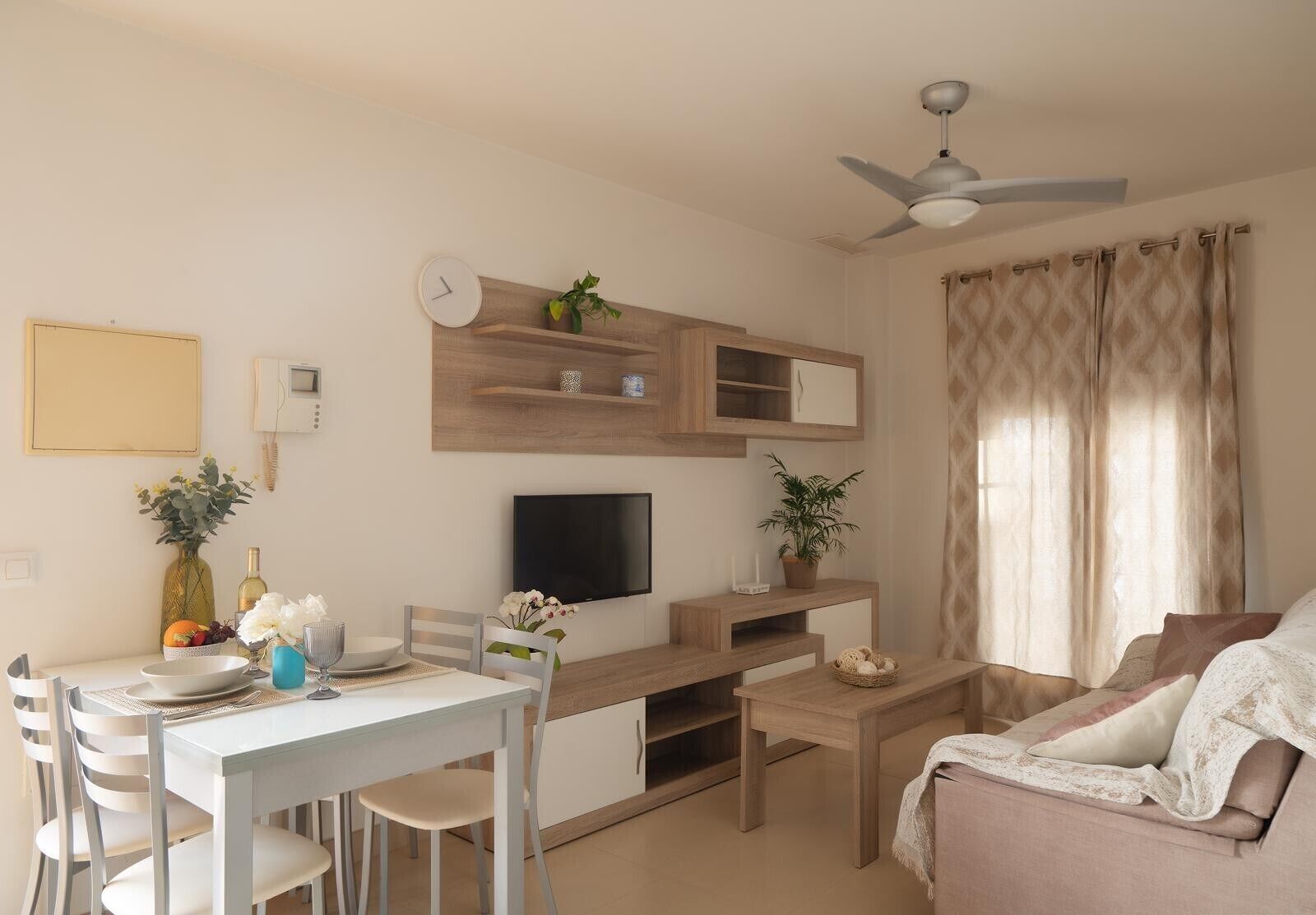 Un salon moderne comprenant un coin repas, un téléviseur à écran plat, des étagères en bois, un ventilateur de plafond et une grande fenêtre avec des rideaux.