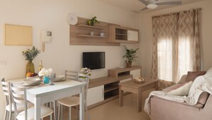 A modern living room with a dining area, a flat-screen TV, wooden shelving, a ceiling fan, and a large window with curtains. - Apartment in the City Center (Conil de la Frontera)