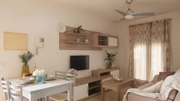 Un salon moderne comprenant un coin repas, un téléviseur à écran plat, des étagères en bois, un ventilateur de plafond et une grande fenêtre avec des rideaux.