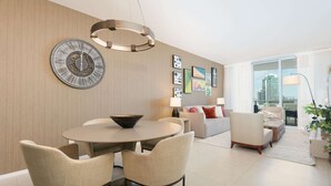 A modern living room with a dining area, featuring a round table, chairs, a large wall clock, and artwork. - 1BR Beach Condo | Sea View | Hollywood Broadwalk (Hollywood)