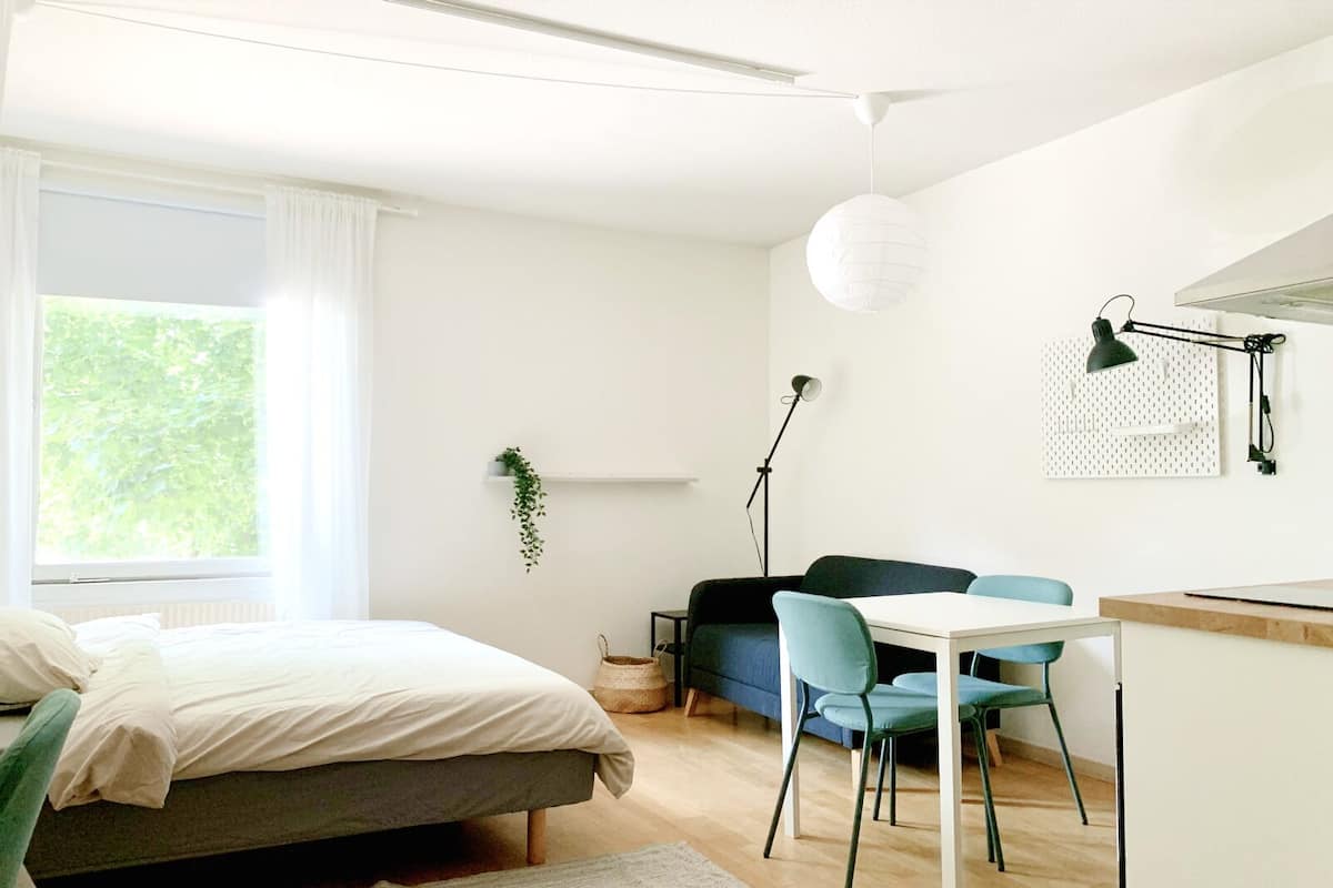 Ein modernes Schlafzimmer mit einem Bett, einem Schreibtisch und einem Stuhl. Ein Fenster mit Vorhängen, eine hängende Pflanze und eine Stehlampe sind ebenfalls vorhanden.