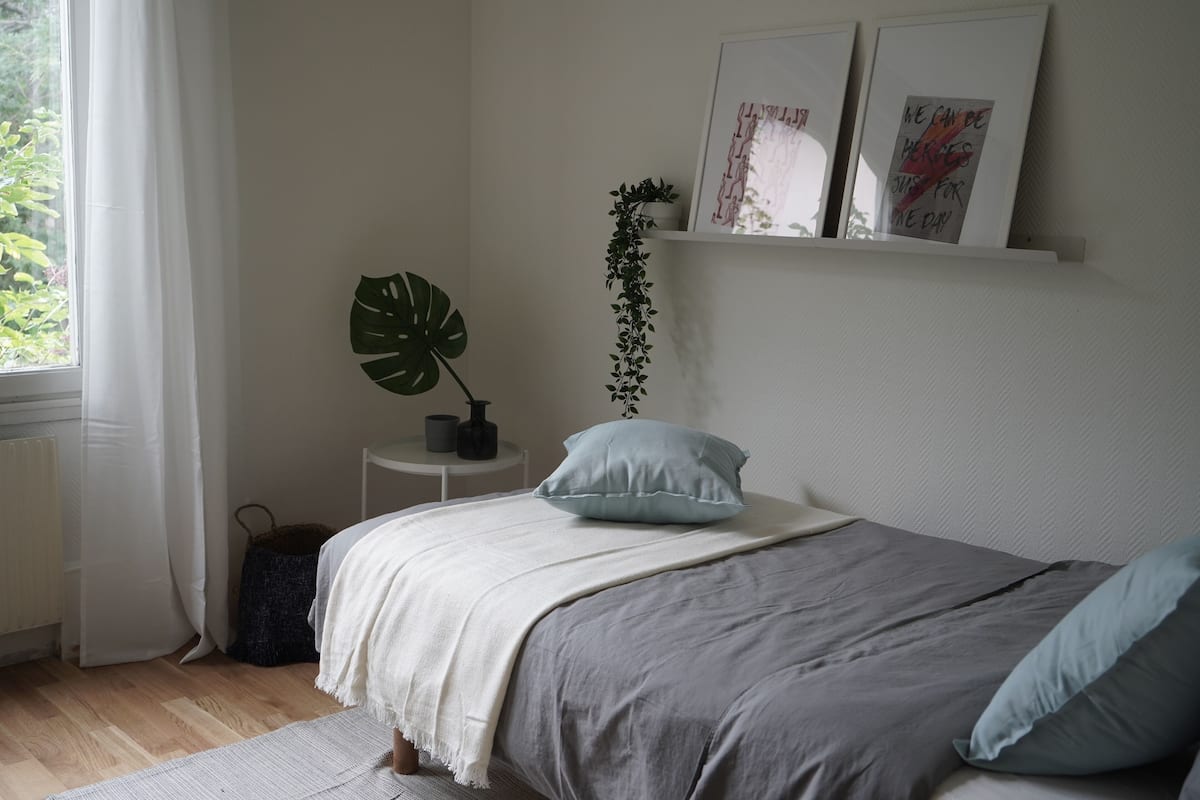 Ein Schlafzimmer mit einem Bett, einem Fenster mit Vorhängen, einer Pflanze und einem kleinen Tisch mit einer Lampe.