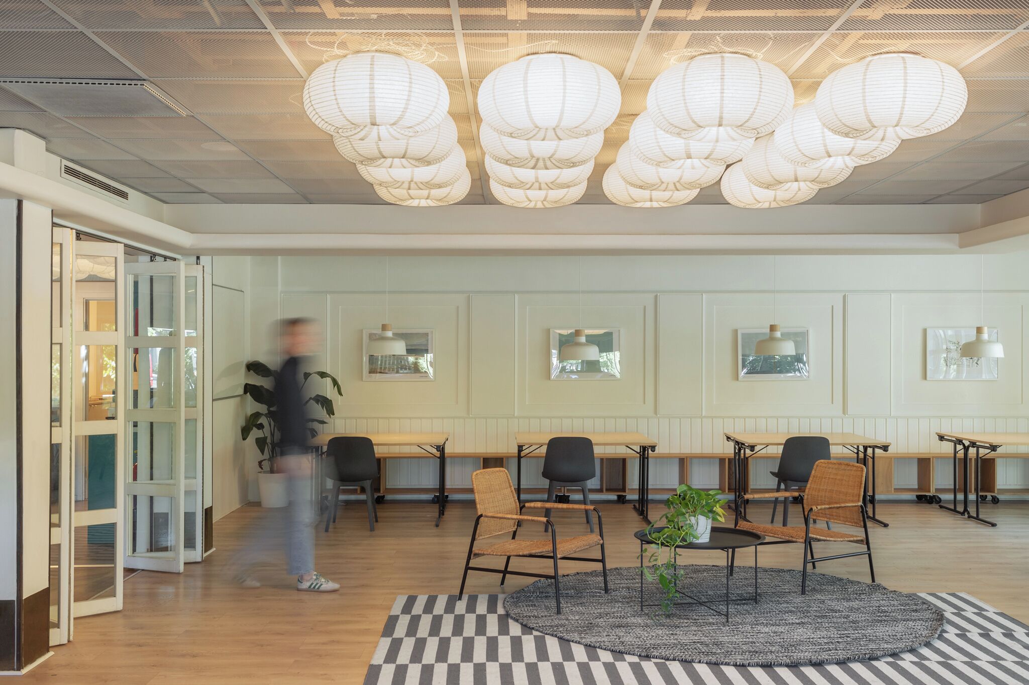 A modern lobby with wooden tables and chairs, a checkered rug, and decorative paper lanterns on the ceiling.