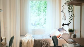 A cozy bedroom with a bed, a desk, and a chair. There is a window with curtains, a plant, and a wall-mounted shelf. - Allihoop - Bromma Friends (Bromma)