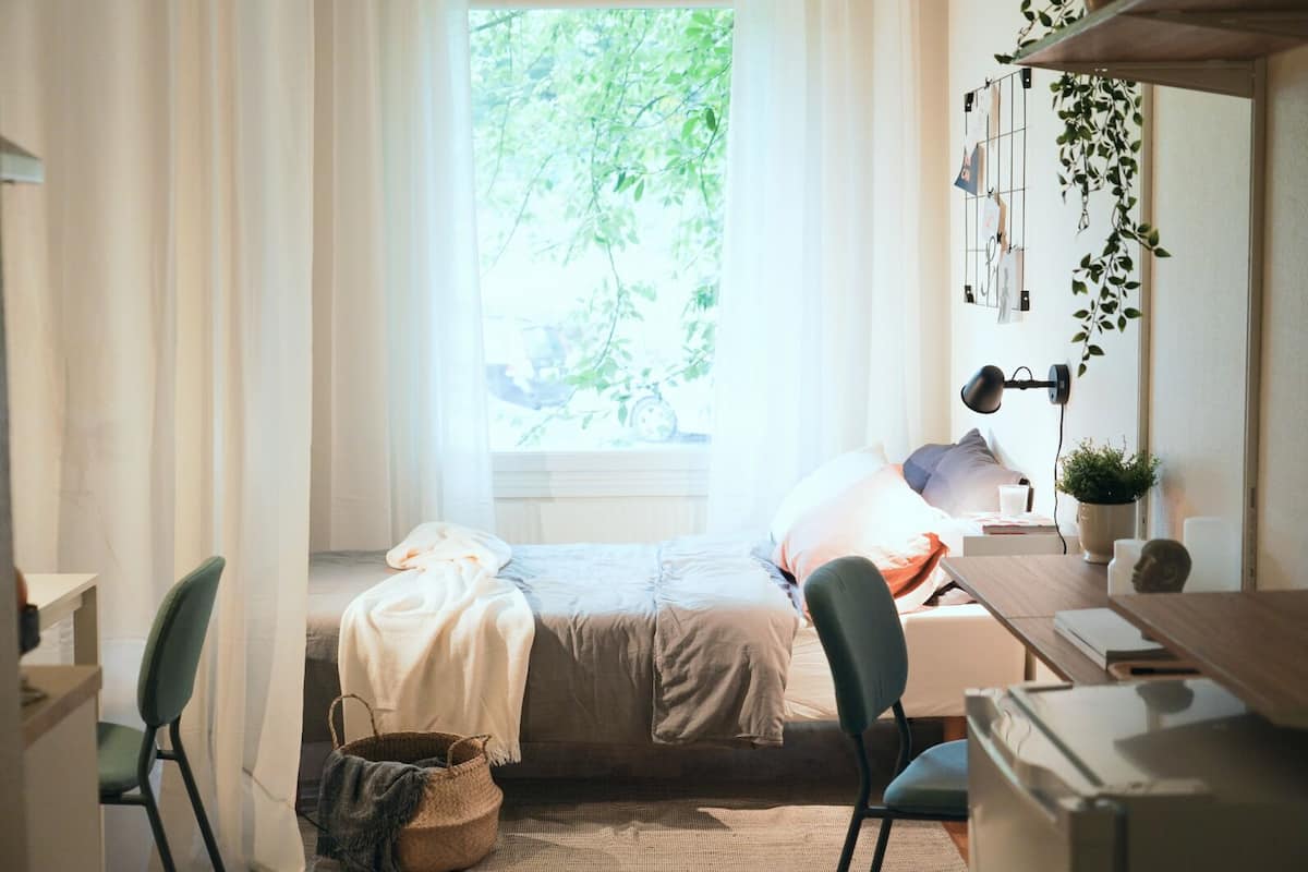 Ein gemütliches Schlafzimmer mit einem Bett, einem Schreibtisch und einem Stuhl. Es gibt ein Fenster mit Vorhängen, eine Pflanze und ein an der Wand befestigtes Regal.