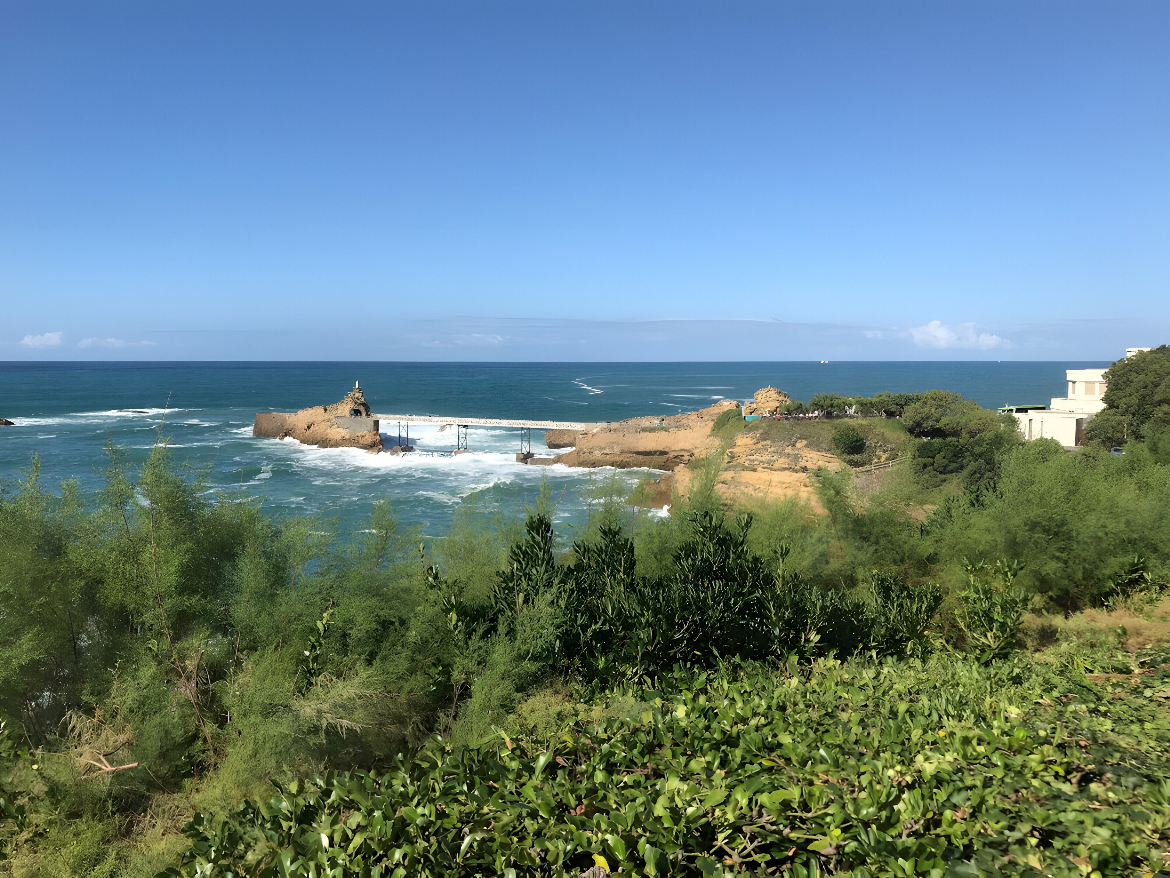 A coastal landscape with a rocky outcrop, a pier, and dense greenery.
