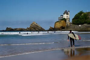 Un paysage côtier avec un bâtiment ressemblant à un château perché sur une falaise, des vagues et des surfeurs.