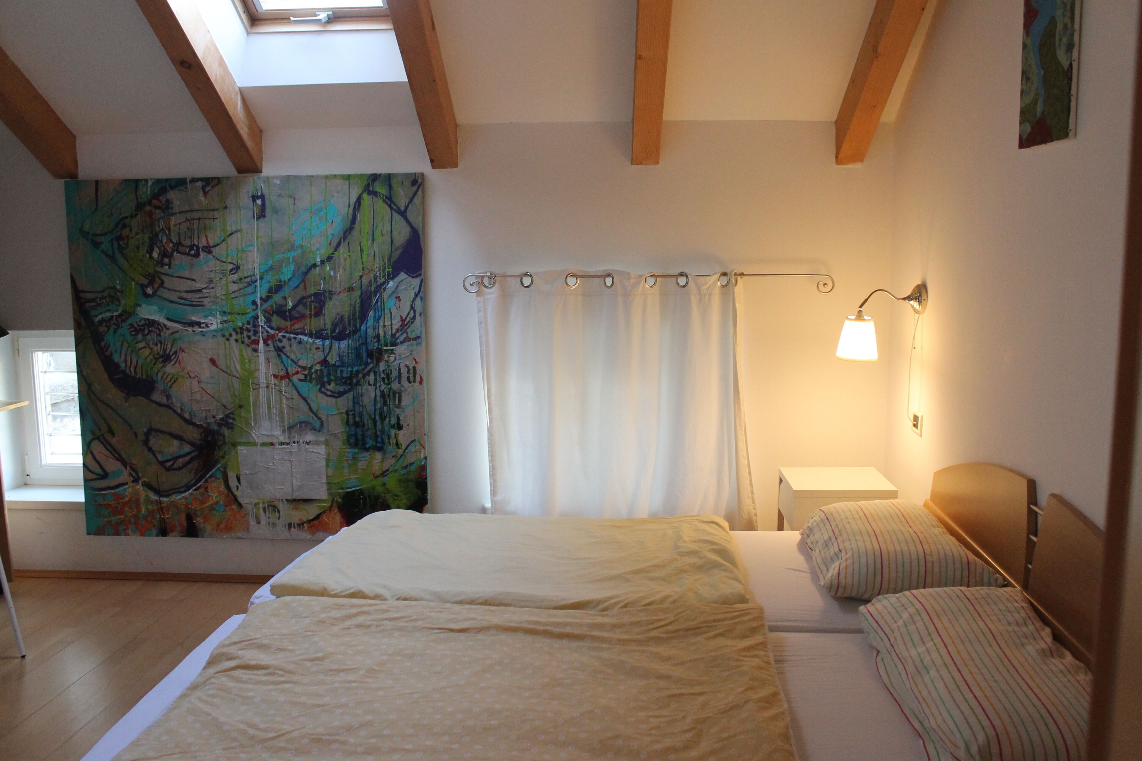 A bedroom with a large bed, a bedside table, a lamp, a window, and a painting on the wall.