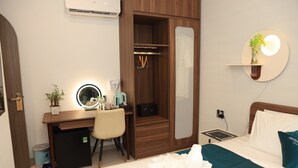 Ein Hotelzimmer mit Bett, Schreibtisch mit Stuhl, kleinem Kühlschrank, Nachttisch mit Lampe, Wandregal mit Pflanze und Duschbereich.