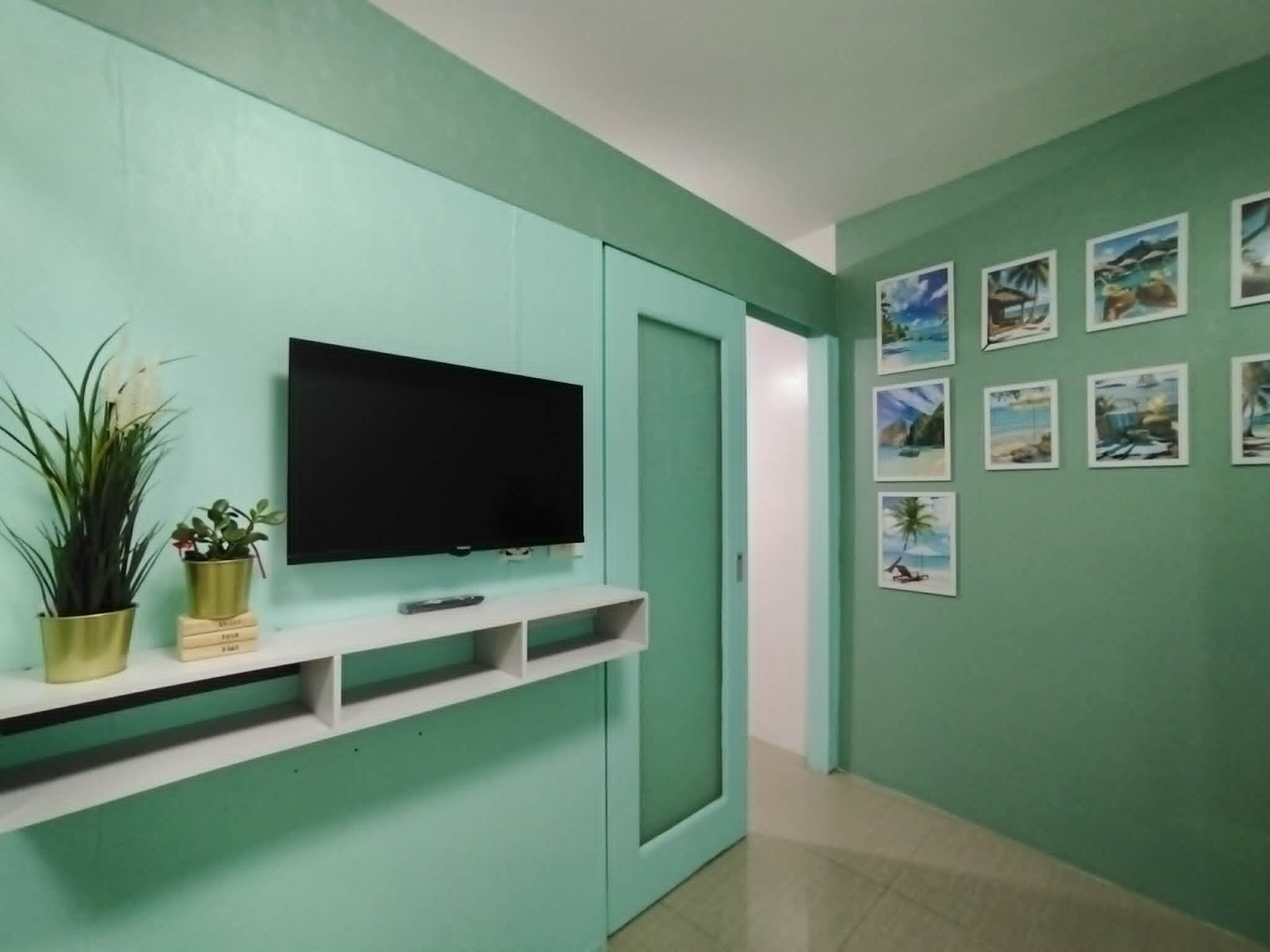 A teal-colored room with a mounted TV, a white shelf with decorative items, and a door leading to another room.