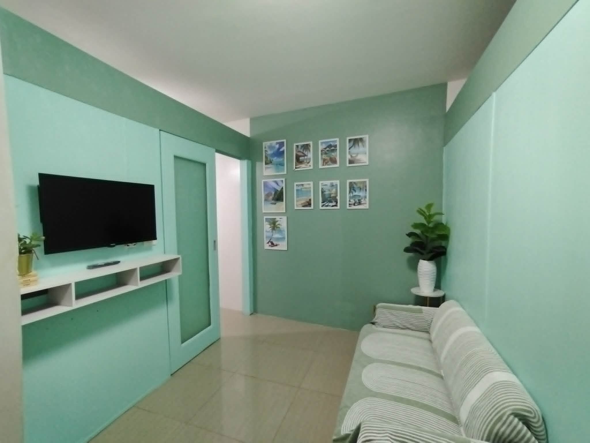 Sala de estar com paredes verde-água, uma televisão de tela plana presa a um armário branco, um sofá e um vaso de plantas.