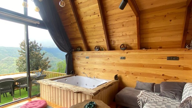 Una cabaña acogedora con interior de madera, un ventanal con vistas panorámicas, un jacuzzi y un espacio de descanso confortable.