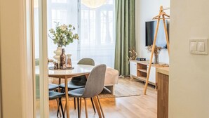 En moderne stue med spiseplads, med et træspisebord, grå polstrede stole, et stort vindue med grønne gardiner, en hængende lampe og et fjernsyn på et træstel.