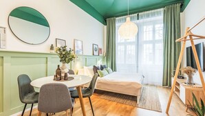 Et moderne værelse med et rundt spisebord, to stole, en seng med grøn gavl, en hængende lysekrone og en gyngestol i træ.
