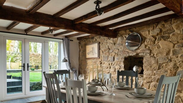 Un espace repas agrémenté d’un mur en pierre, de poutres en bois et offrant une vue sur le jardin grâce à des portes vitrées.