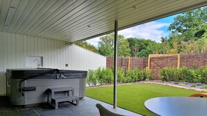 Eine überdachte Terrasse mit integriertem Whirlpool, einem runden Tisch und Blick auf einen gepflegten Garten und Bäume.