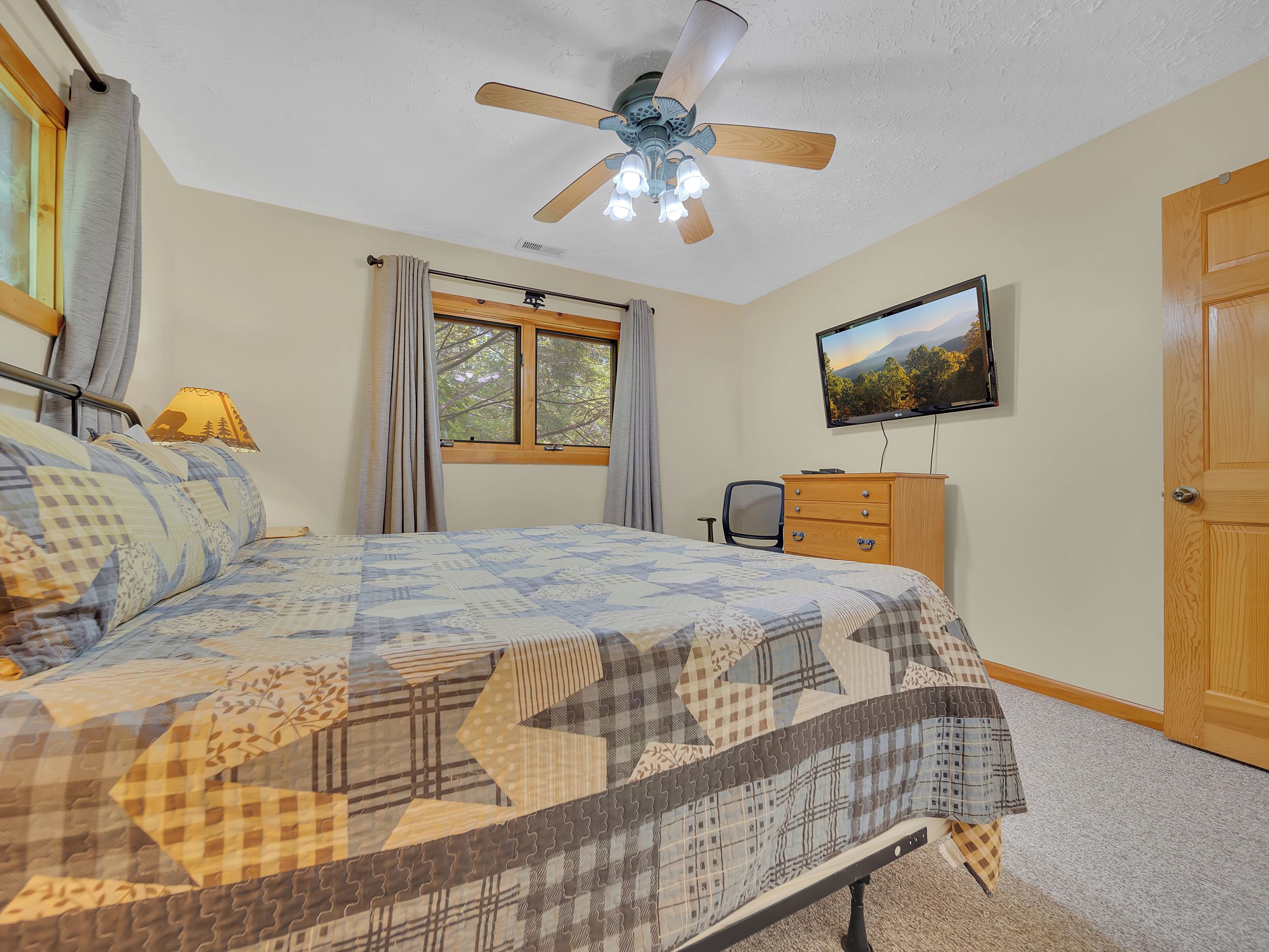 Un dormitorio con cama, ventilador de techo, ventana con cortinas, televisor en la pared y cómoda de madera.