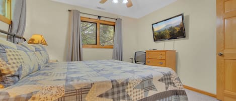Un dormitorio con cama, ventilador de techo, ventana con cortinas, televisor en la pared y cómoda de madera.