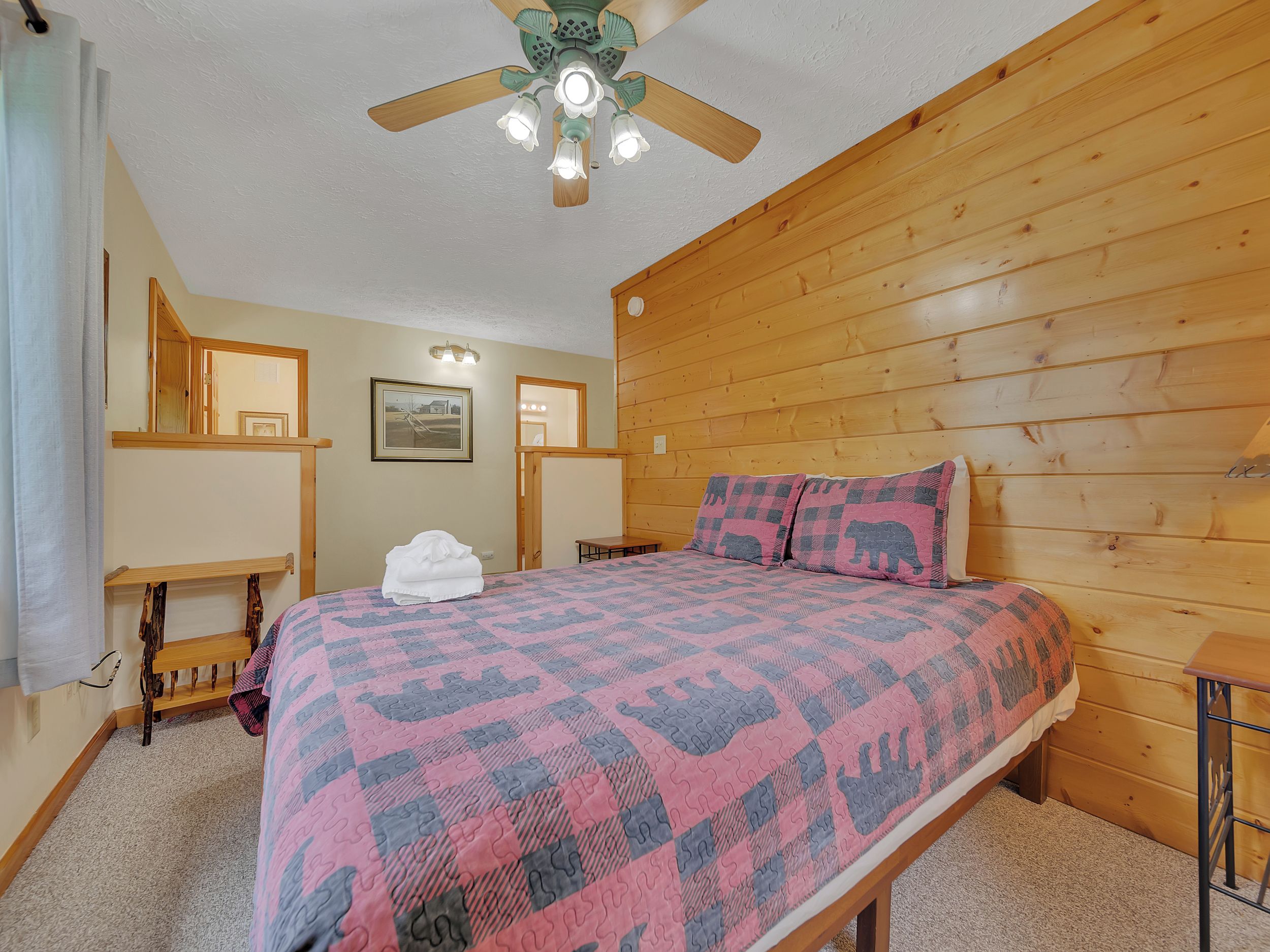Un dormitorio con cabecera de madera, cama con colcha estampada, ventilador de techo y una pequeña mesa con silla.