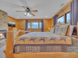 Un dormitorio acogedor de cabaña de madera con cama, chimenea, ventilador de techo y un ventanal con vistas al exterior.