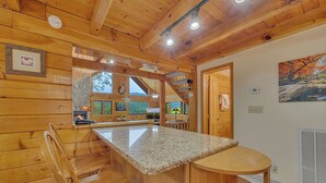 A wooden cabin interior with a kitchen area featuring a granite countertop, wooden chairs, and a stone fireplace.