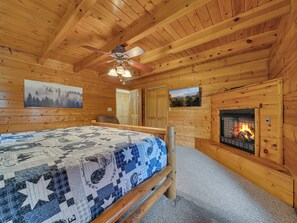 A cozy log cabin room with a bed, a fireplace, a ceiling fan, and two wall-mounted televisions. - Cabin w/Great View|Hot Tub|Pool Table & Air Hockey (Gatlinburg)