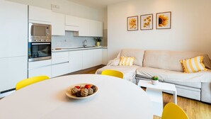 Une cuisine moderne avec des armoires blanches, un four encastré et un four à micro-ondes. On y trouve une table à manger avec des chaises jaunes et un saladier rempli de fruits. Un canapé avec des coussins jaunes et blancs se trouve dans l’espace salon.