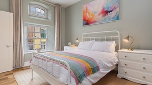 Een slaapkamer met een bed, nachtkastjes, een dressoir, een raam met gordijnen en een kleurrijk abstract schilderij aan de muur.