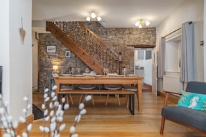 Un coin repas avec une table en bois dressée pour quatre personnes, un mur en pierre, un escalier en bois et une vue sur une chambre à coucher par une porte ouverte.