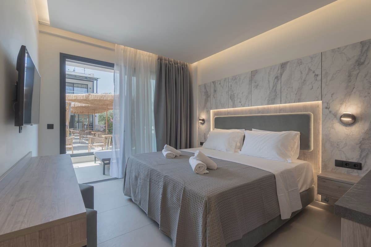 A modern hotel room with a large bed, a flat-screen TV, a wooden desk, and a balcony with outdoor seating.