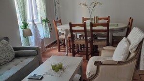 A cozy living room with a sofa, a coffee table with remote controls and glasses, a dining table with chairs, and a potted plant.