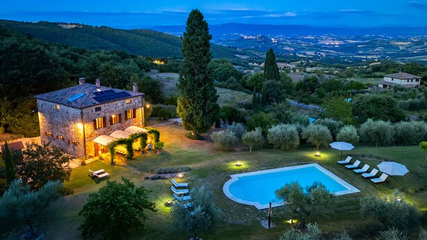 A stone villa with a swimming pool, surrounded by lush greenery and a view of distant hills.