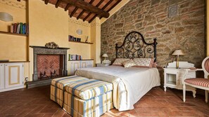 A bedroom with a stone wall, a fireplace, a bed with a striped ottoman, a nightstand, and a chair.