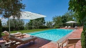 A rectangular swimming pool with clear blue water, surrounded by a tiled patio. There are wooden lounge chairs and white umbrellas providing shade.