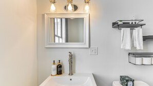 Een badkamer met een witte wastafel, een spiegel en een toilet. Twee flessen zeep en lotion, een pak toiletpapier en een plank met handdoeken.