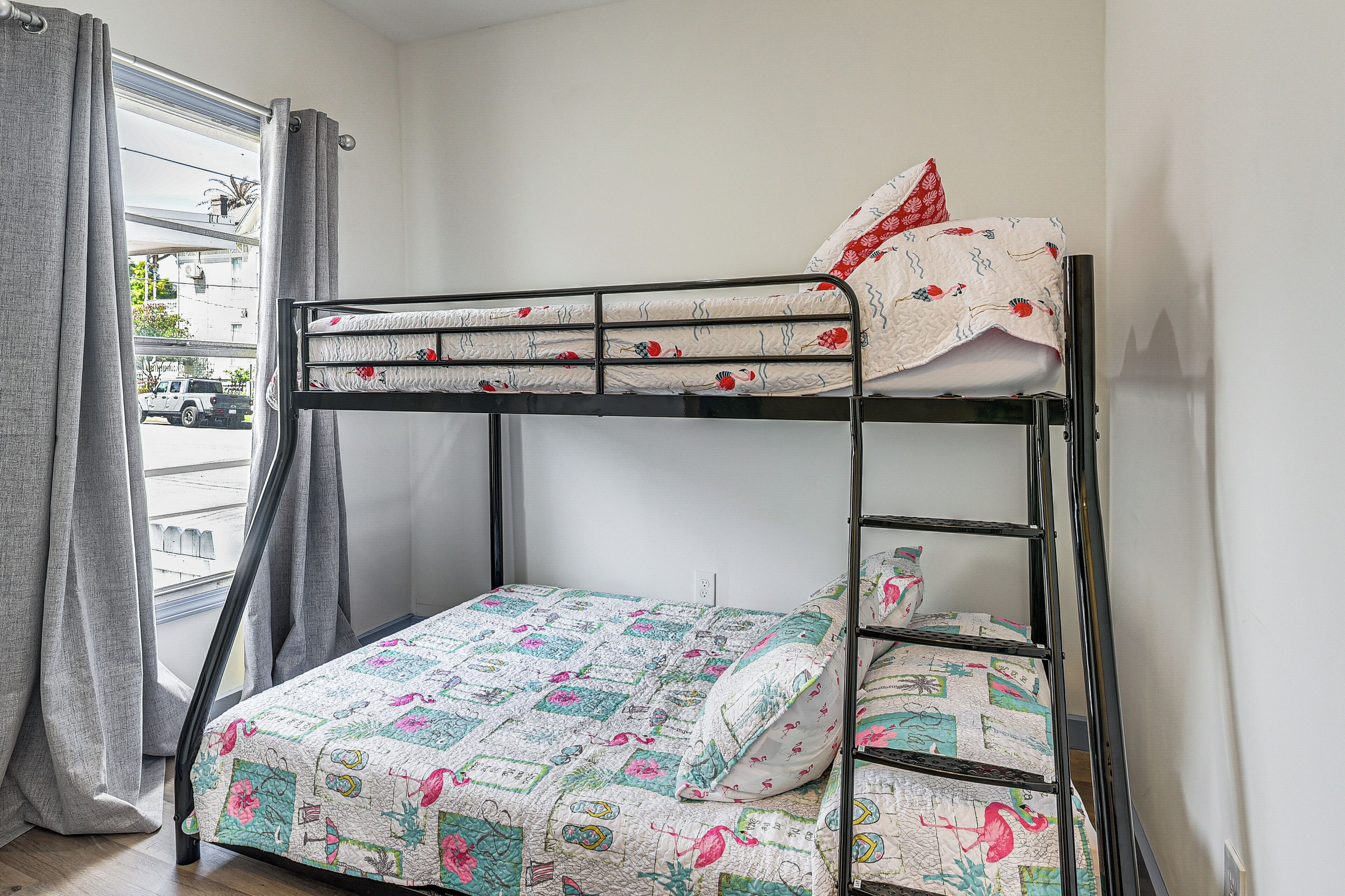 Una cama litera con colcha y almohadas estampadas, una ventana con cortinas, y vista a la calle.