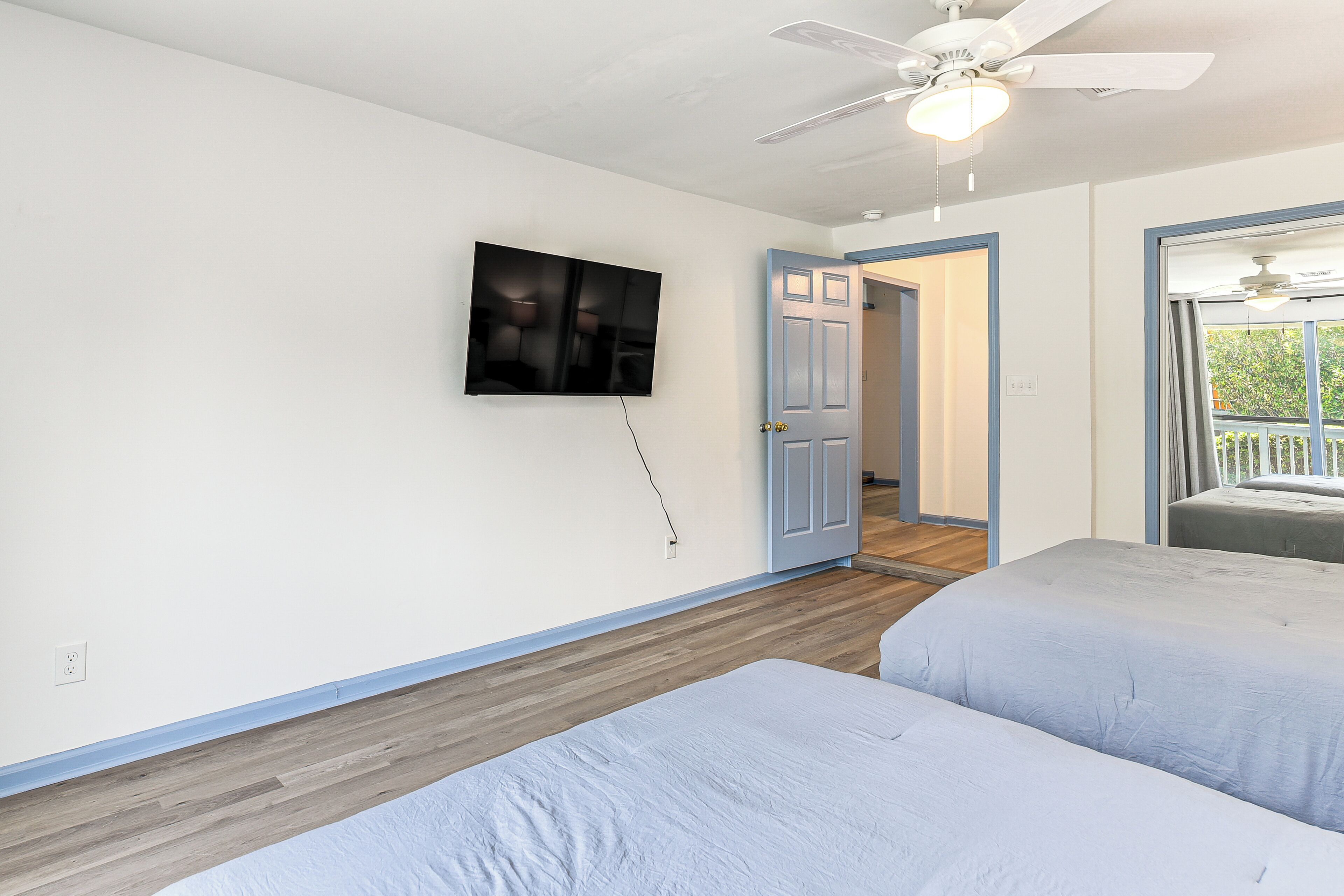 A bedroom with two beds, a ceiling fan, a TV mounted on the wall, and a door leading to another room.