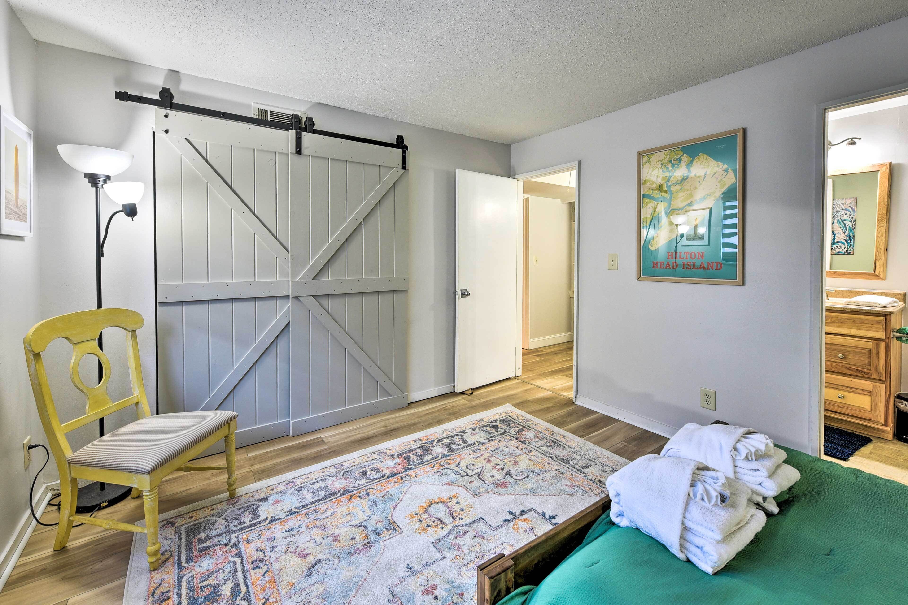 Een slaapkamer met een grote schuifdeur in boerderijstijl, een stoel, een kamerkleed met een patroon, een bed met een groene spreideken en een schilderij van Hilton Head Island.