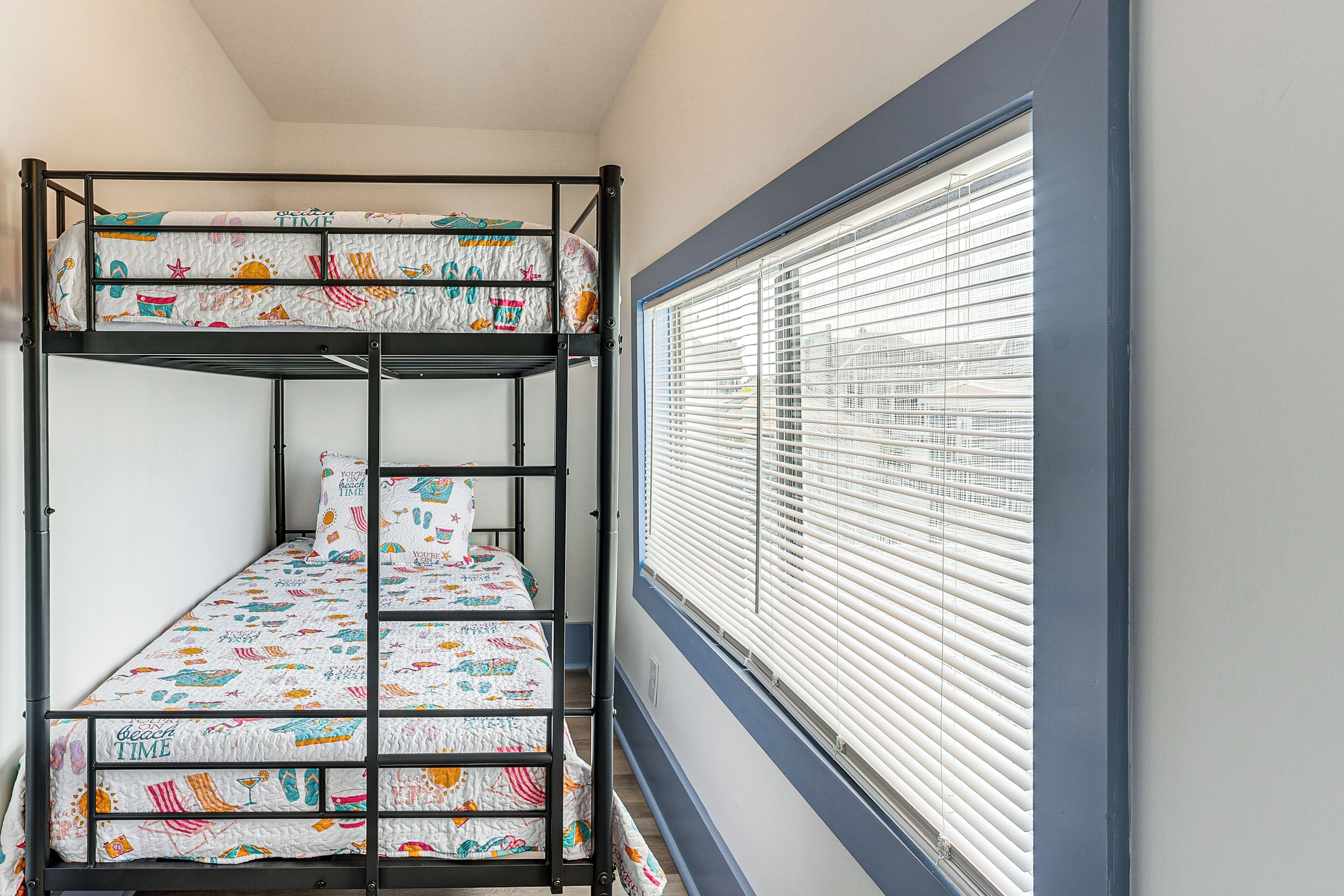 Una cama nido con ropa de cama temática de playa, una ventana con persianas y una pared lisa.