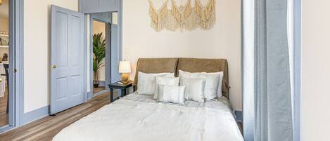 A bedroom with a large bed, a nightstand with a lamp, and a macrame wall hanging.