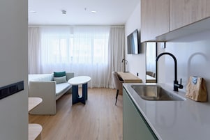 A modern kitchen with a sink, a dining area with a table and chairs, a sofa, and a TV mounted on the wall.