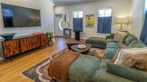 A living room with a large flat-screen TV, a wooden cabinet, a green sofa, a coffee table, and a patterned rug. - Bold, Fun Getaway near Lake, Zoo, Downtown (Michigan City)