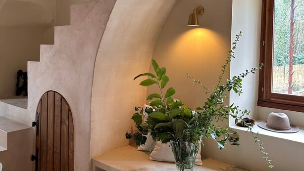 Una escalera con techo de vigas de madera, un ventanal con vistas a árboles y un florero con plantas sobre un repisa blanca.