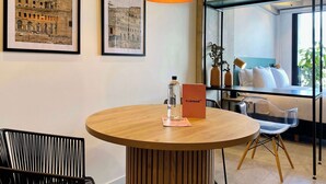 A modern interior with a round wooden table, framed pictures on the wall, a hanging pendant light, and a view of the outdoors through a window.