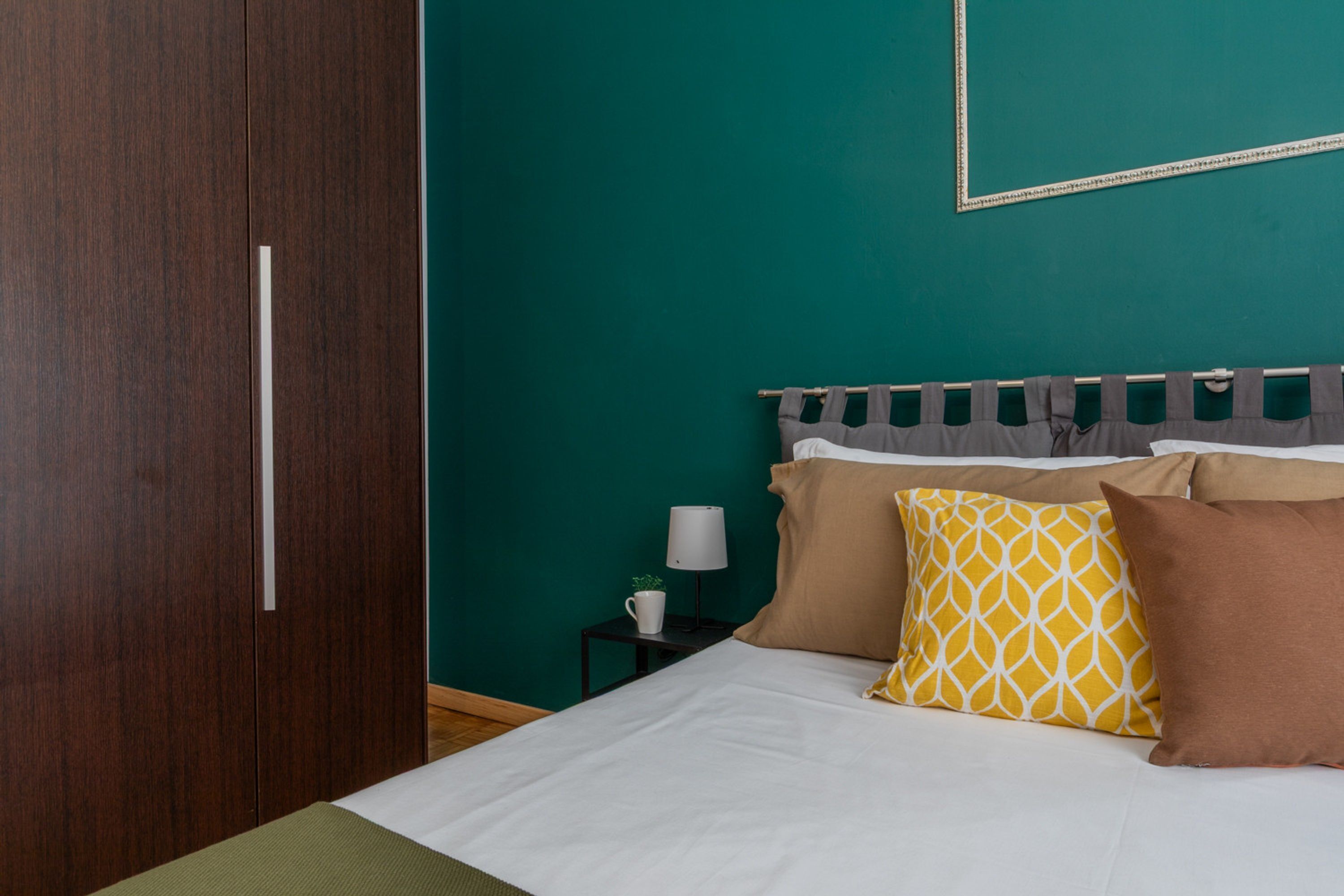 Een netjes opgemaakt bed met een bruin-witte hoofdbord, een klein nachtkastje met een lamp en een donkere houten garderobe.