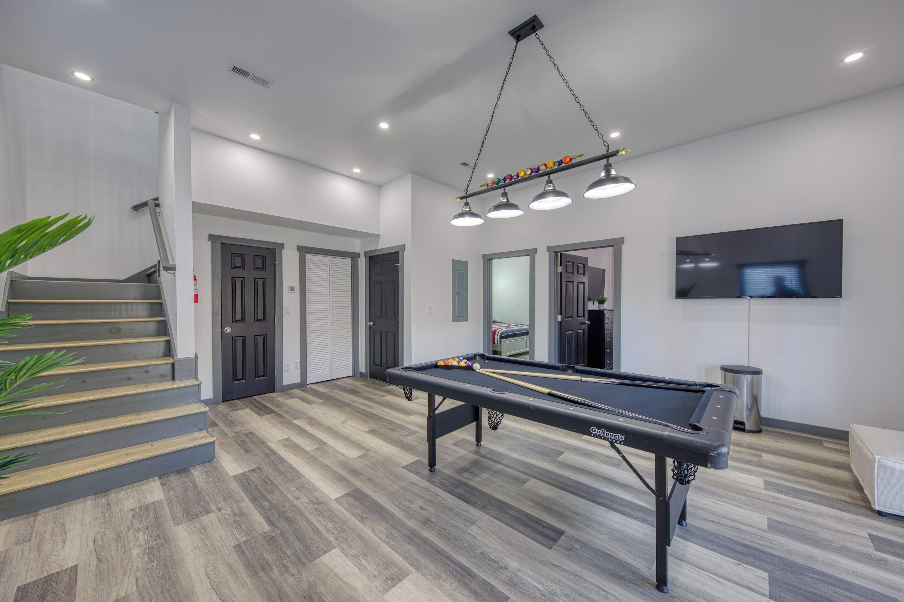 A modern indoor pool table with overhead lighting, a staircase, and a television on the wall.