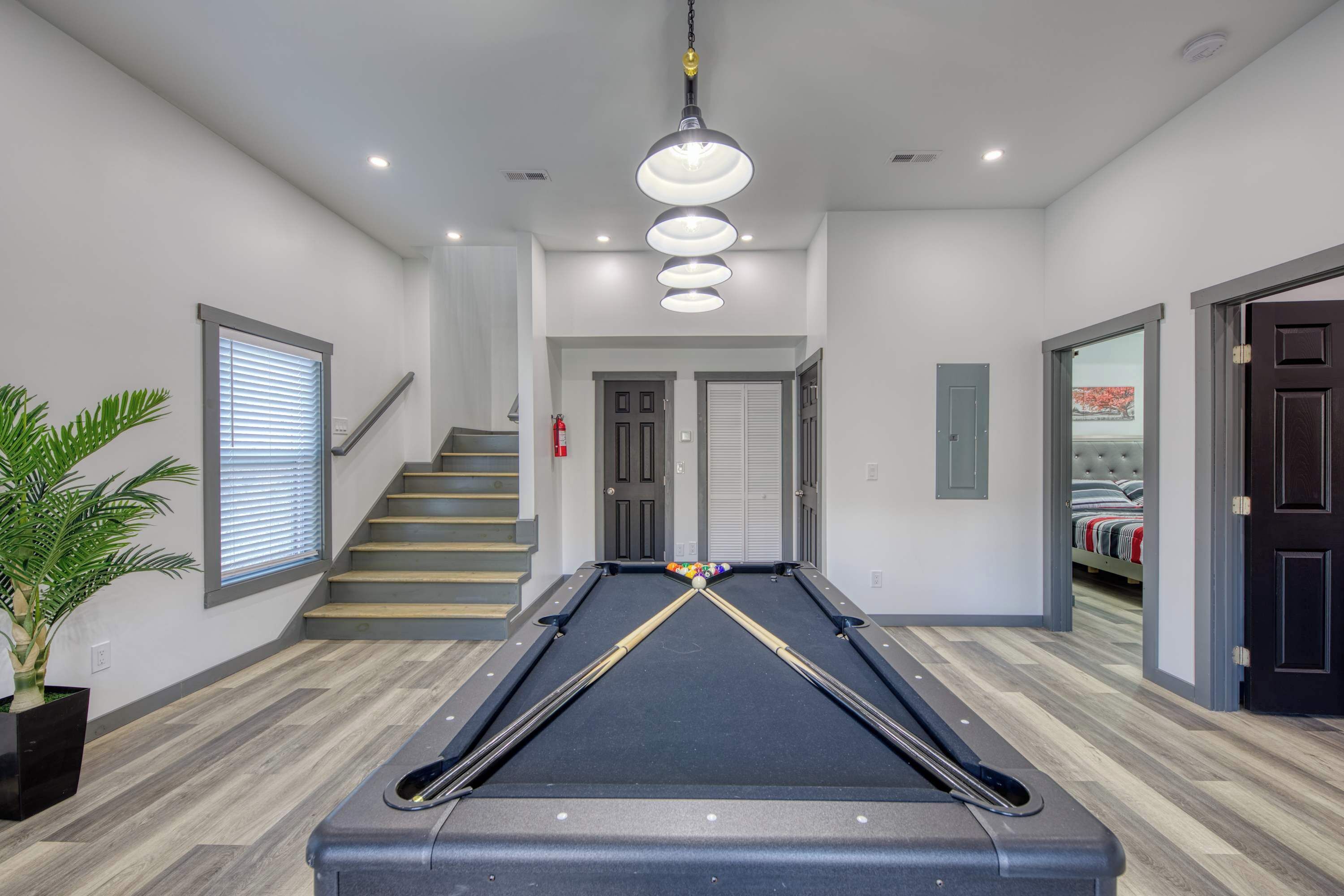 A pool table in a modern room with a staircase, a potted plant, and a bed visible through an open door.