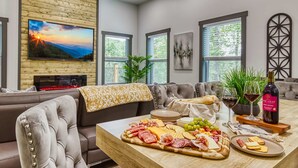 A modern living room with a large TV, a wooden wall panel, a sofa, a dining table with a variety of cheeses and meats, and a bottle of wine. - Bearfoot Getaway - New Lux Cabin Hot Tub Pool (Gatlinburg)